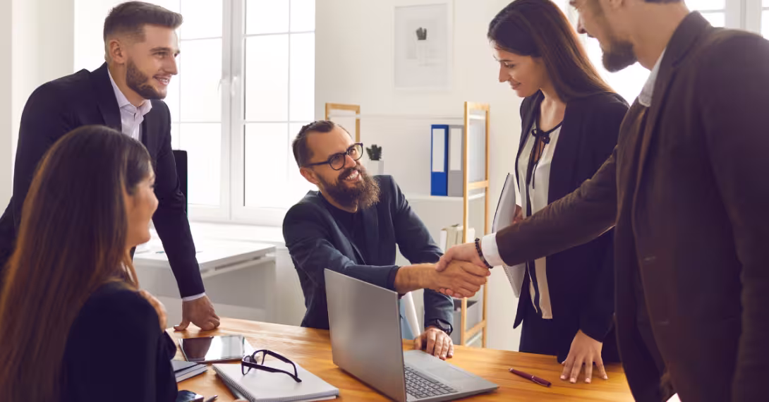 Five people around a desk with a laptop on it, one man shaking the hand of another